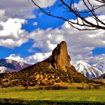 large rock formation shaped like a needle, with snow capped mountains in the back ground and green fields in the foreground
