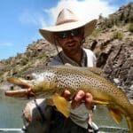 Black Canyon Anglers man in a white hat holding a spotted trout fish with a rugged canyon wall in the background