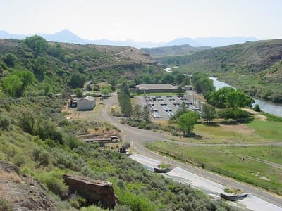 A wide-angle view of the Hitchkiss Fish Hatchery situated in a dry, green river canyon. The complex includes a main building and long, rectangular outdoor fish raceways, visible next to a winding, blue-green river. The facility is surrounded by arid, brush-covered hillsides and is set against a distant backdrop of low mountains under a clear, bright sky.
