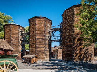 Three large, hexagonal wooden grain silos connected by walkways in a historic pioneer town setting under a clear blue sky.