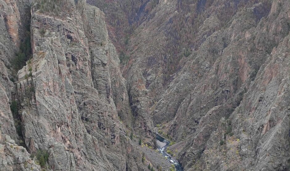 A dramatic, vertical view looking deep into the Black Canyon of the Gunnison. The steep, sheer walls of dark Precambrian rock drop to a winding river far below under an overcast sky.