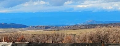 A wide view over ranch corrals and a fence in the foreground. Beyond the fence, a vast, dry plain leads to distant rolling hills and mountains under a bright, cloudy sky with a band of vivid blue color.