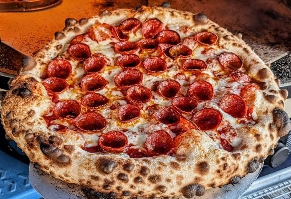 Close-up of a large, perfectly baked pepperoni pizza, featuring a naturally leavened, dark, bubbly crust and "cup-and-char" pepperoni slices over melted mozzarella cheese.