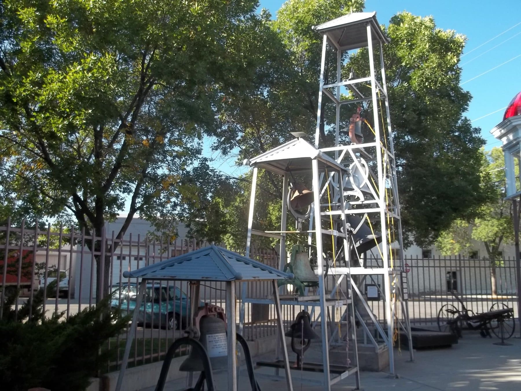 "A photograph of an outdoor bell display at the Grand Mesa Arts & Events Center. Several large, vintage-style bells of various sizes and colors are mounted within tall, silver-painted metal frame towers. The display is situated on a paved area, enclosed by a black metal fence, with large green trees and a clear blue sky in the background. A small wooden structure with a gabled roof sits in the foreground, housing one of the bells."
