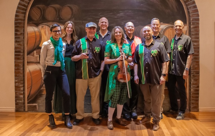 Promotional photo and event details for the Sinners & Saints St. Patrick's Concert. The photo shows a nine-member band posing in front of a mural of wine barrels. Several members wear black shirts with green vertical stripes and shamrock icons; one woman in the center wears a green 18th-century style frock coat and holds a fiddle