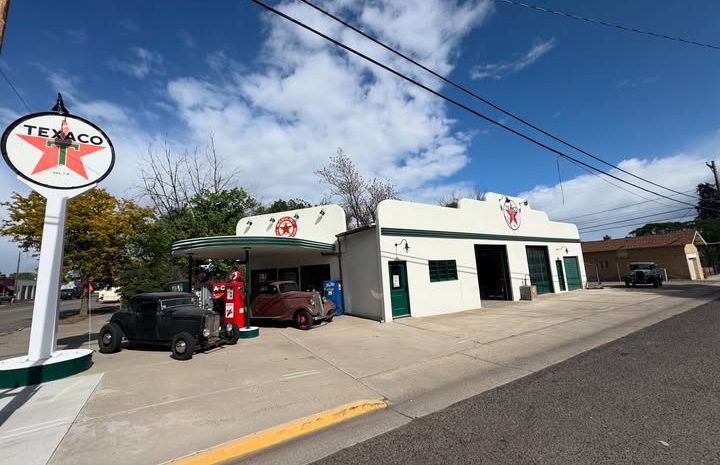 A pristine white vintage Texaco service station in Hotchkiss, Colorado, with classic cars parked under the bay. Clear blue sky and retro gas pumps visible.