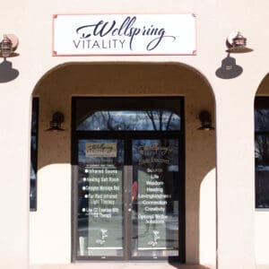 Building Exterior and Entrance
The exterior entrance of Wellspring Vitality, featuring a light-colored stucco building with three large architectural arches. A white rectangular sign above the center arch displays the "Wellspring Vitality" logo in purple script. The glass front doors are adorned with text listing available therapies, including Infrared Sauna, Healing Salt Room, and Red Light Therapy.