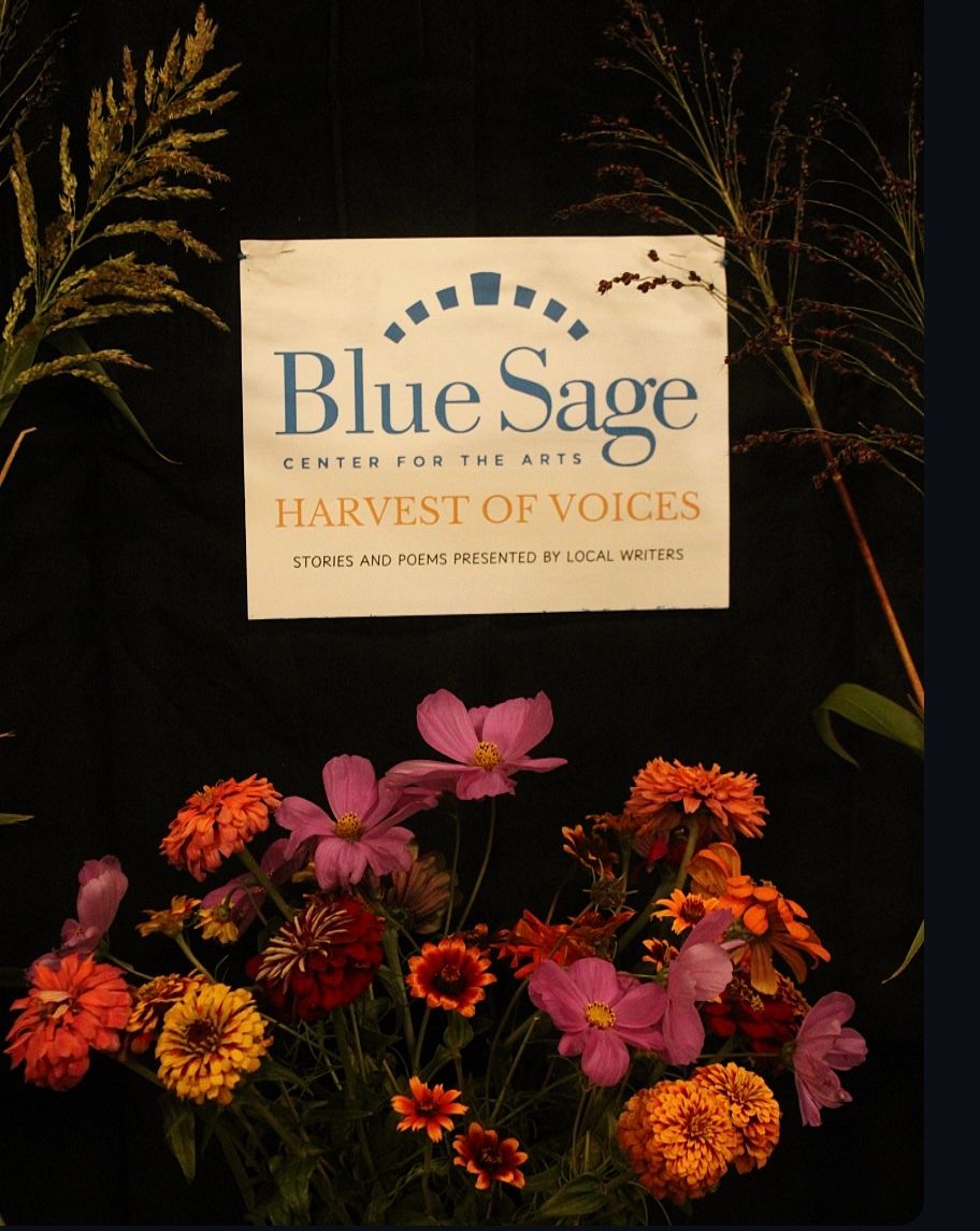 A white sign on a dark background reads "Blue Sage Center for the Arts Harvest of Voices." Colorful fall flowers, including orange and pink zinnias, bloom beneath the sign.