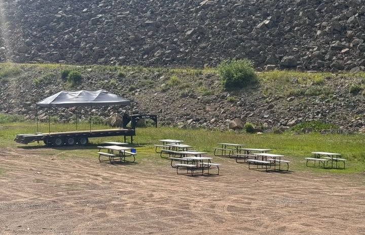 An outdoor event area at Grand Mesa Lodge featuring rows of white picnic tables positioned on a dirt lot in front of a flatbed trailer stage under a canopy tent, backed by a large rocky hillside.