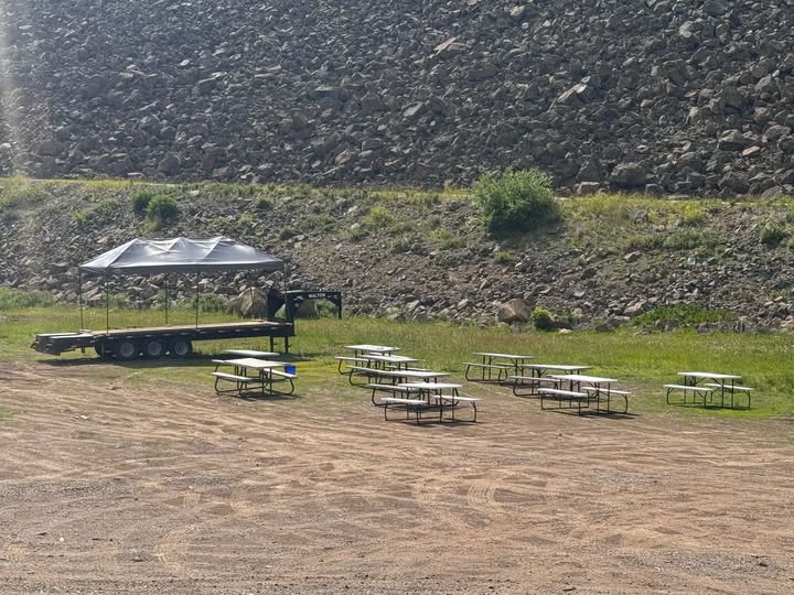 An outdoor event area at Grand Mesa Lodge featuring rows of white picnic tables positioned on a dirt lot in front of a flatbed trailer stage under a canopy tent, backed by a large rocky hillside.