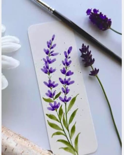 Close-up photo of a hand-painted watercolor bookmark featuring delicate purple lavender stems and green leaves resting on a white table next to paintbrushes and real lavender sprigs.
