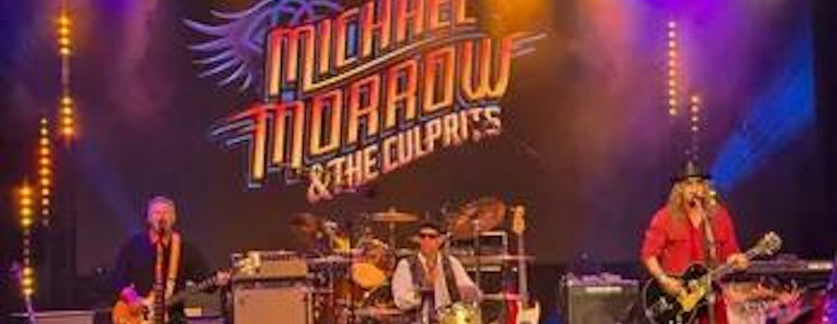 Live stage photo of three-piece band Michael Morrow & The Culprits performing under vibrant blue and yellow stage lights with a large band logo projected behind them.